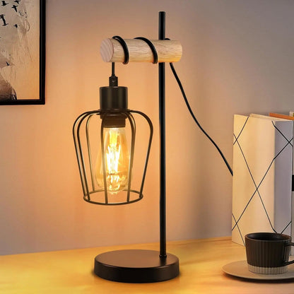 Lampe à poser industrielle avec abat-jour cage en métal noir et potence en bois clair allumée sur un bureau à côté de livres